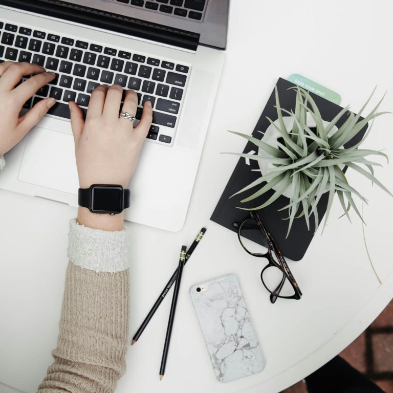 Virtual Assistant. Amber Assists. Person typing on laptop with sage colored plant beside them. glasses. apple watch.